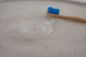 Set of clear aligner trays on a gray background with a wooden toothbrush with blue bristles blurry in the background